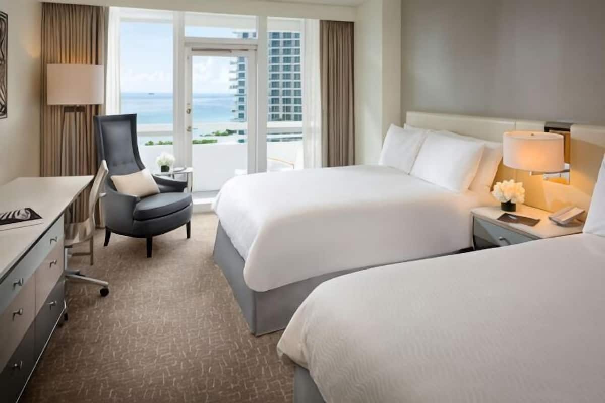 A hotel room with two beds, a desk, a chair, a lamp, and a view of the ocean.