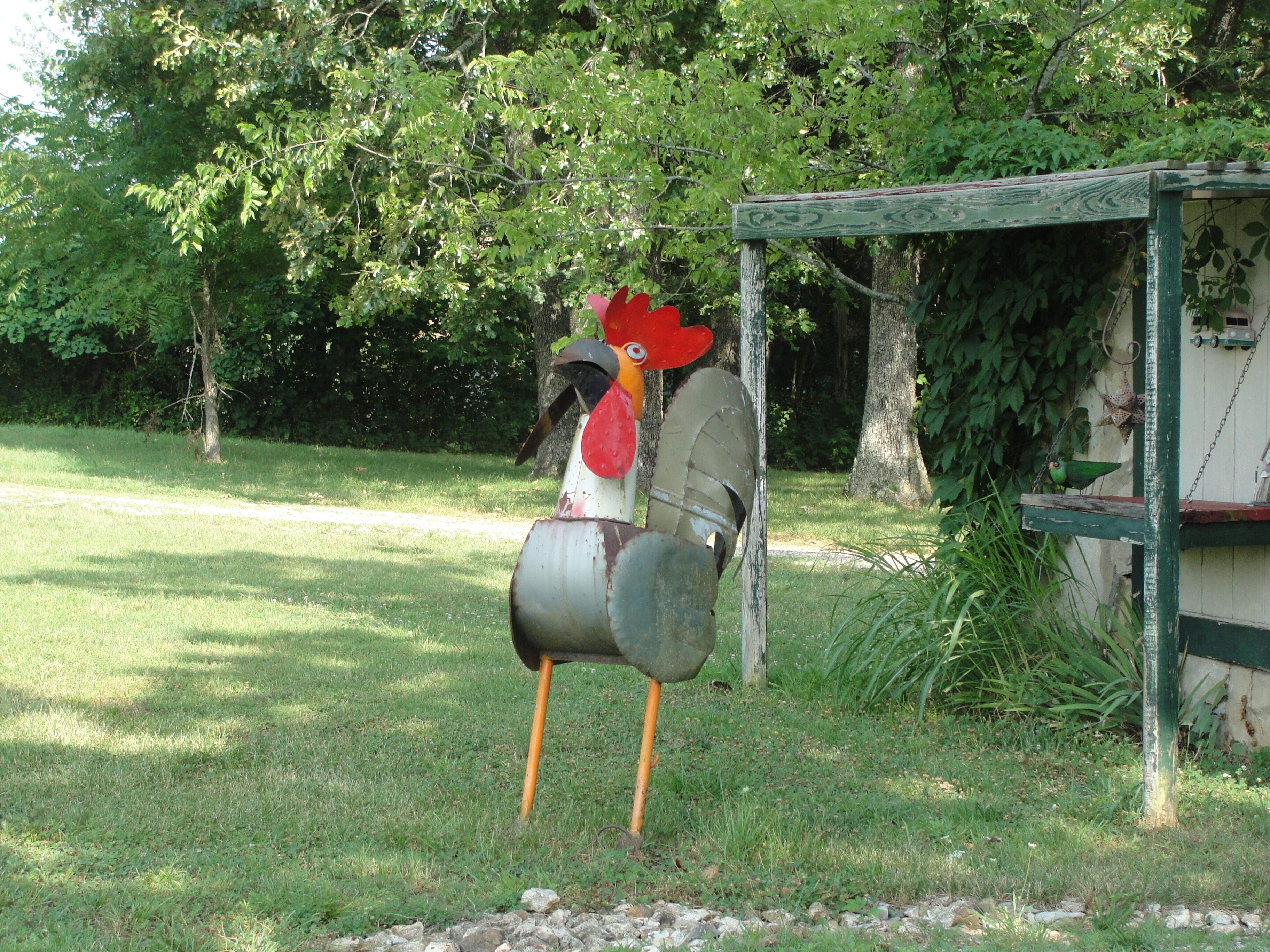 Un cadre extérieur rustique agrémenté d’une sculpture de coq décorative, placée sur un socle et entourée de verdure et d’arbres.