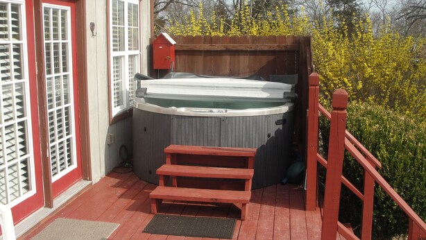 A hot tub on a wooden deck with a red railing, a wooden house with red trim, and yellow flowering bushes in the background. - Stone Meadow Resort (Eureka Springs)