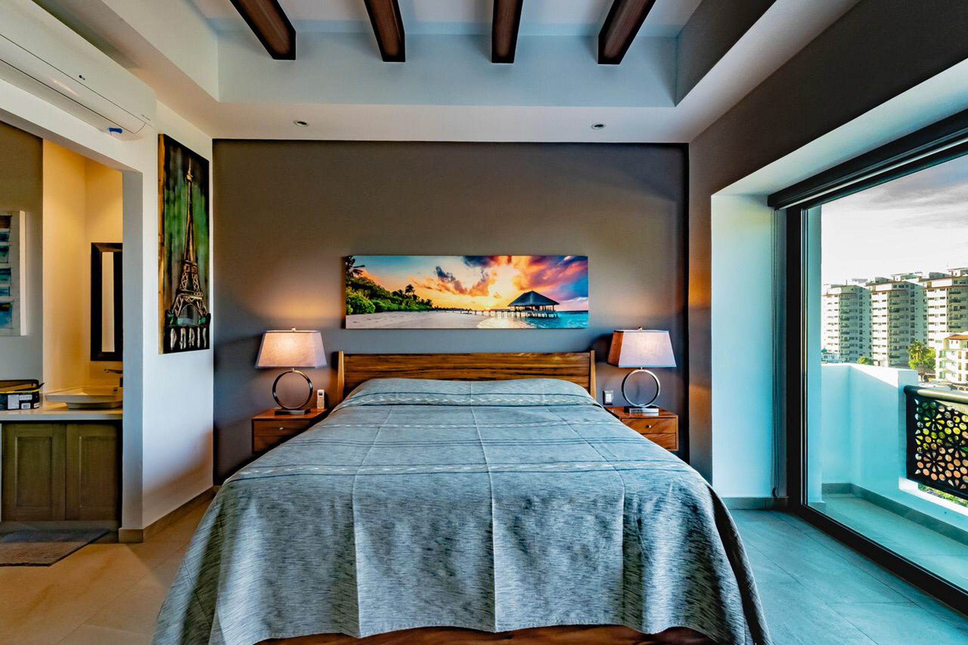 Een moderne slaapkamer met een groot bed, een schilderij van een zonsondergang en een balkon met uitzicht op gebouwen.