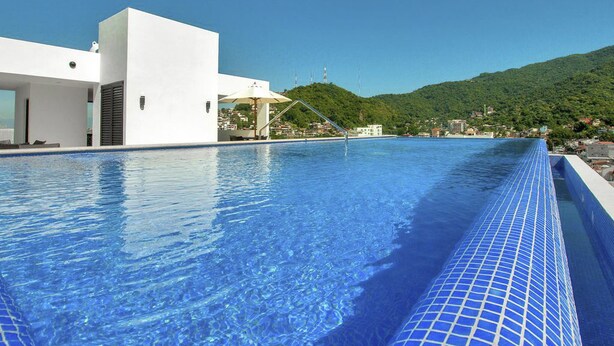 A modern hotel with a long swimming pool, a white building, and a view of a green hillside. - Casa Iguana at Carranza (Puerto Vallarta)