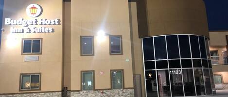 Un hotel con diseño moderno llamado Budget Host Inn & Suites, que destaca por su entrada de cristal curvo y numerosas ventanas.