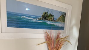 Un tableau représentant un paysage côtier, encadré, est accroché au-dessus d’une table en bois sur laquelle se trouve un vase rempli d’herbes séchées.