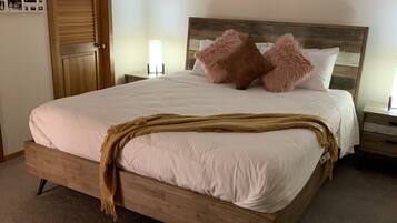 A bedroom with a wooden bed, a door, a wall-mounted picture frame, and a bedside table with a lamp.