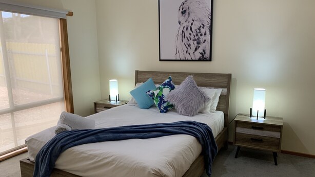Une chambre à coucher comprenant un lit, deux tables de chevet, une commode, une grande fenêtre et une œuvre d’art accrochée au mur.