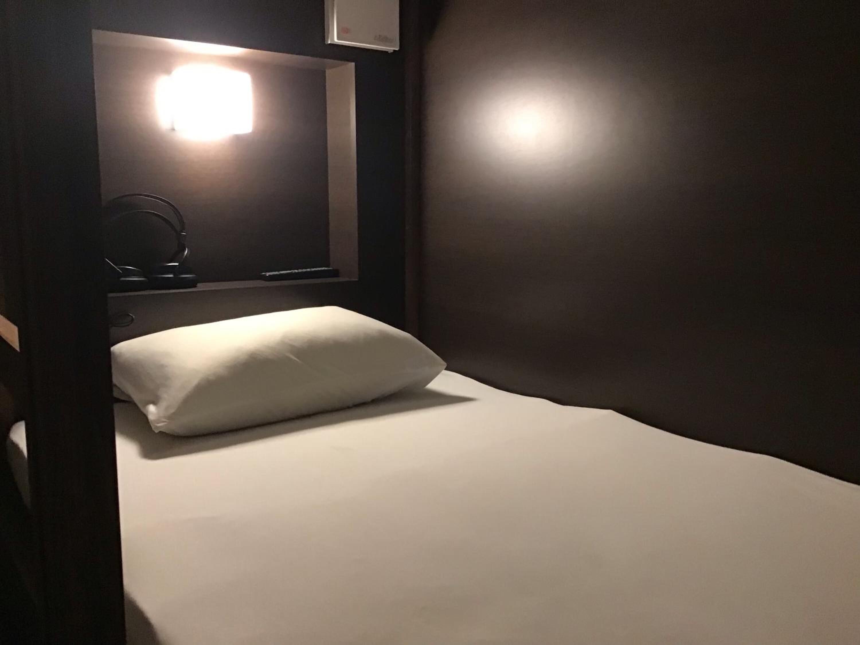 Una habitación tenuemente iluminada con una cama individual, un almohadón blanco y una mesita con una lámpara.