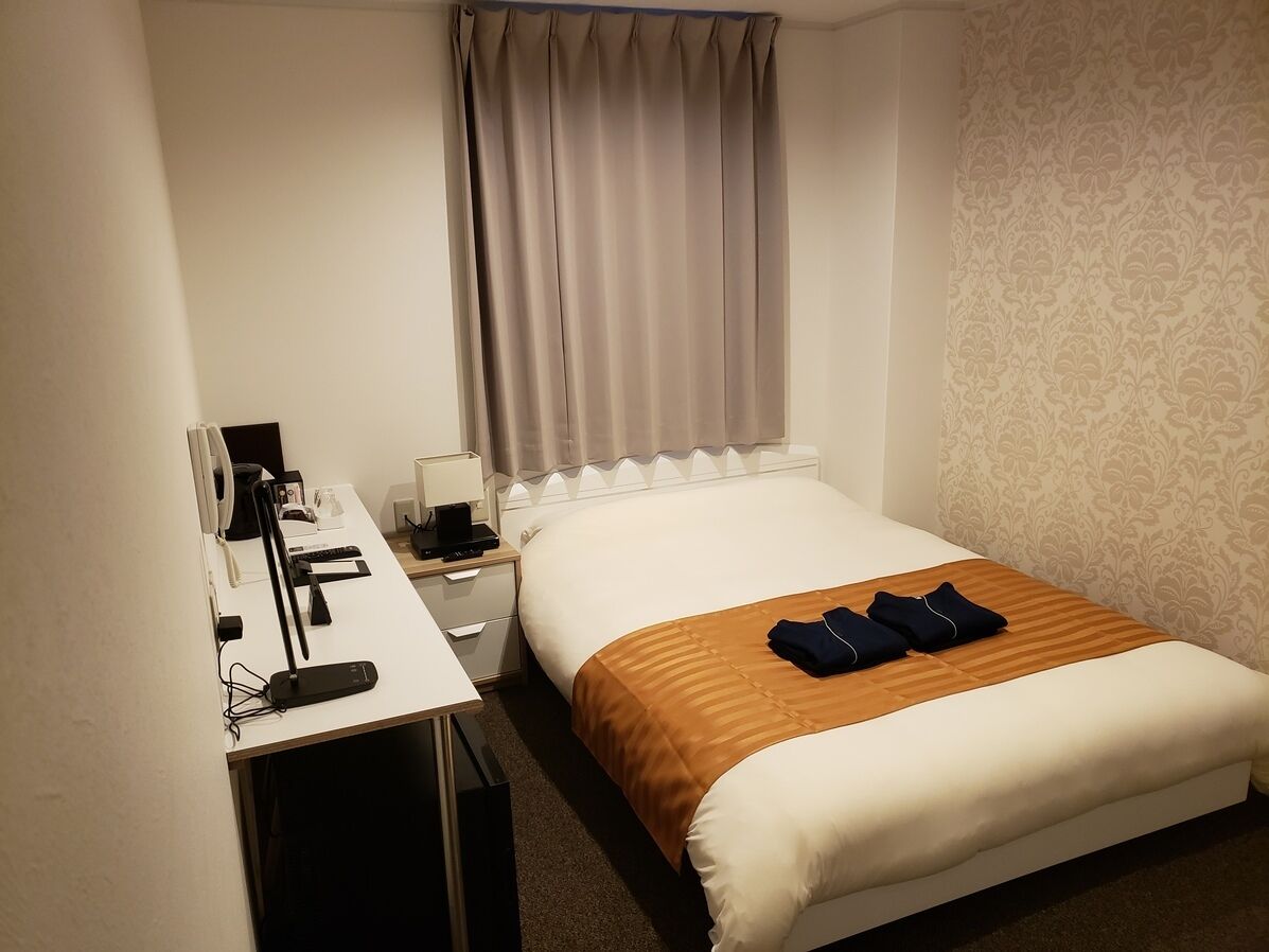 A hotel room with a bed, a desk with a lamp, a chair, a window with curtains, and a patterned wall.
