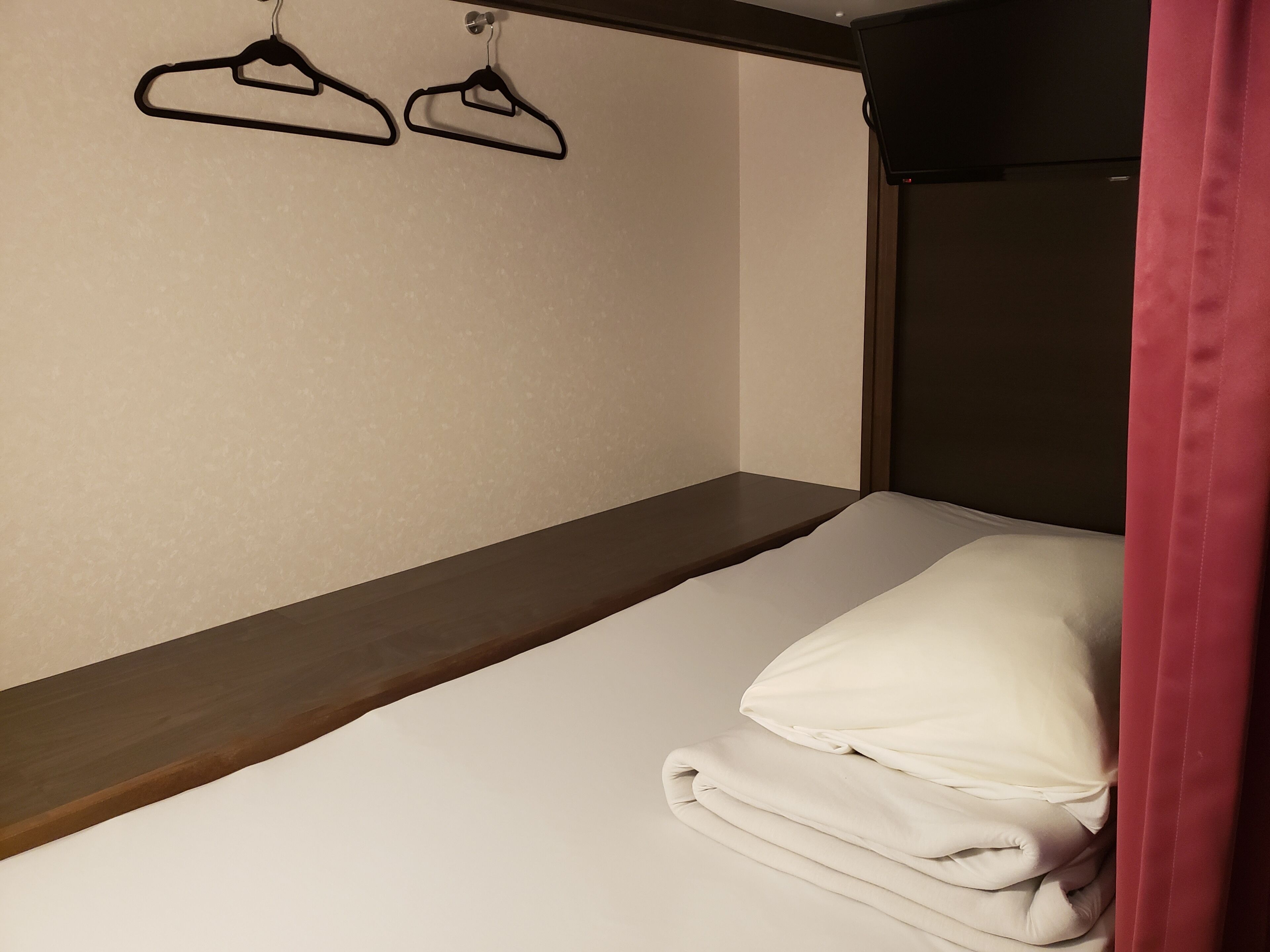 A single bed with a wooden headboard, a white mattress, and a folded blanket. Two black hangers are visible on the wall above the bed.
