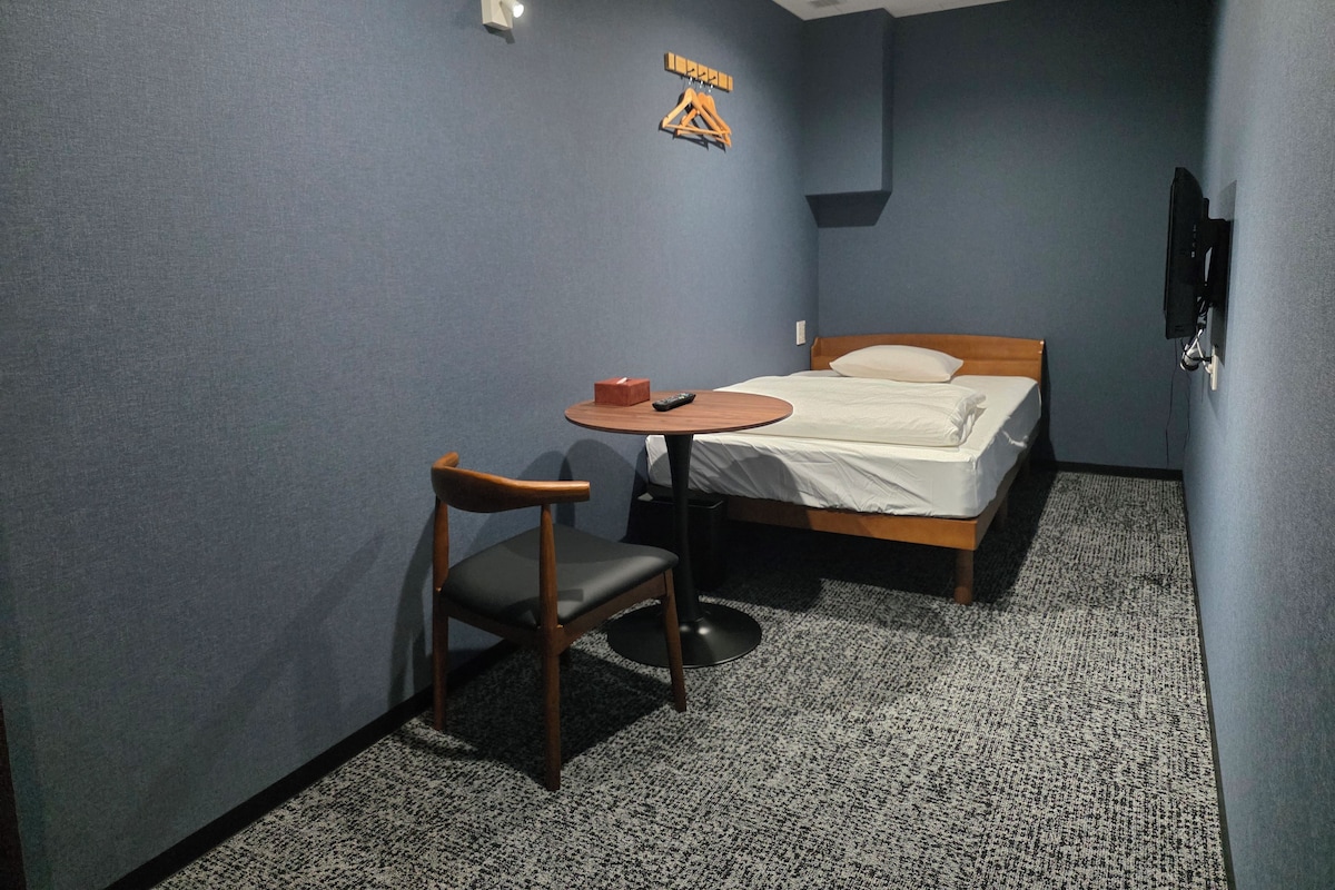 A small room with a bed, a chair, a table, and a wall-mounted TV.