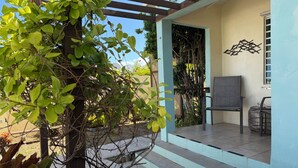 A covered outdoor seating area with a chair and a small table, surrounded by greenery and a wooden pergola. - Coralina- Beautiful Cottage for Two...a Great Value (Vieques)