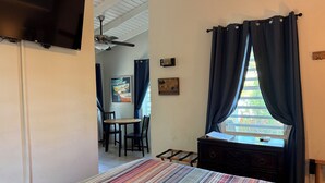 A room with a bed, a window with curtains, a TV mounted on the wall, a ceiling fan, and a small table with chairs. - Coralina- Beautiful Cottage for Two...a Great Value (Vieques)
