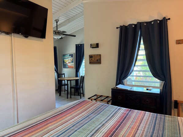 A room with a bed, a window with curtains, a TV mounted on the wall, a ceiling fan, and a small table with chairs. - Coralina- Beautiful Cottage for Two...a Great Value (Vieques)