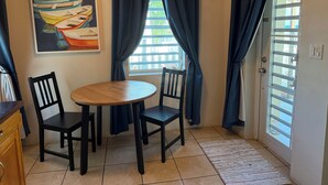 A room with a round wooden table, two black chairs, a painting of boats, and a window with curtains. - Coralina- Beautiful Cottage for Two...a Great Value (Vieques)
