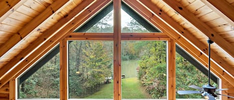 A wooden cabin with a large triangular window offering a view of a lake and surrounding trees.