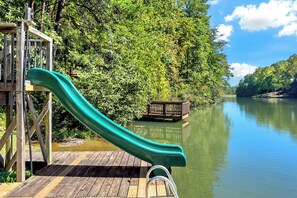 Une terrasse en bois avec un toboggan vert menant à un quai surplombant un lac paisible, le tout entouré d’une végétation luxuriante.