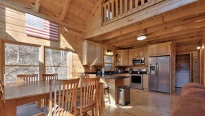 L’intérieur d’un chalet en bois comprend un coin repas, une cuisine et une grande fenêtre donnant sur une forêt.