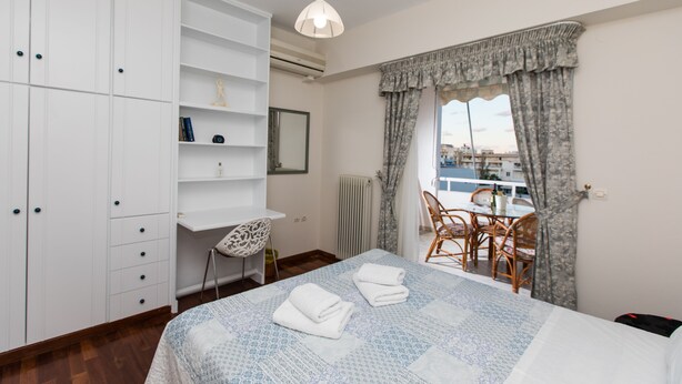 Ein Schlafzimmer mit einem Bett, einem Schreibtisch und einem Stuhl. Ein Fenster mit Vorhängen bietet einen Blick auf Gebäude im Freien.