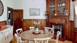 A dining room with a wooden table, white chairs, a fireplace, and a cabinet with glass doors. - Ada Cottage - Warburton, Yarra Valley (Warburton)