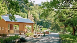 A wooden cabin with a sloped roof, a bench outside, and a paved path leading through a wooded area. - Ada Cottage - Warburton, Yarra Valley (Warburton)