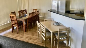 Un coin repas avec des chaises et une table en bois, une cuisine équipée d’un réfrigérateur et d’un évier, et un ventilateur de plafond.