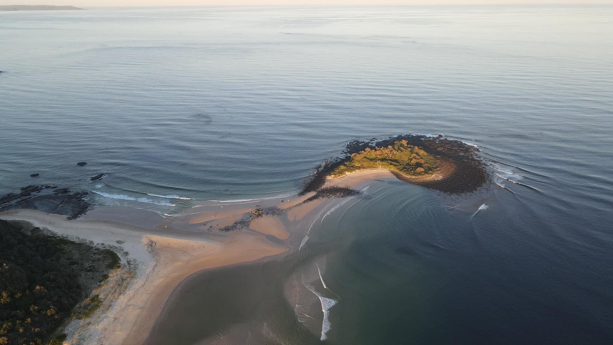 Vue aérienne d’une zone côtière avec une plage de sable, des affleurements rocheux et une petite île couverte de végétation.