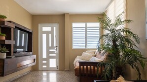 Ein Schlafzimmer mit einem Bett, einem an einer Holzablage montierten Fernseher, einer großen Topfpflanze und einem Fenster mit Jalousien.