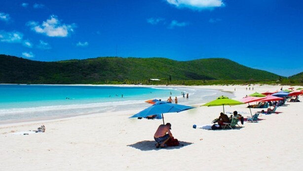 Ein Strand mit weißem Sand, klarem türkisblauem Wasser und grünen Hügeln im Hintergrund. Bunte Sonnenschirme und Menschen entspannen sich am Strand.