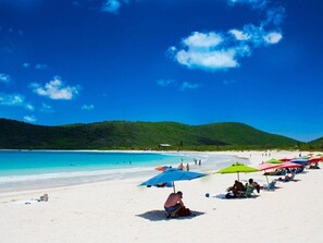 Une plage de sable blanc, une eau turquoise et limpide, et des collines verdoyantes en arrière-plan. On y voit des parasols colorés et des gens qui se détendent sur le sable.