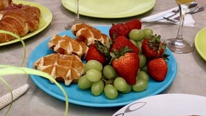 Ein Esstisch mit Tellern voller Speisen, darunter Waffeln, Erdbeeren und Trauben, sowie zwei Gläser Orangensaft, draußen mit einem Pool im Hintergrund.