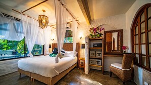 Un dormitorio con una cama grande, dosel, una silla de mimbre, un espejo y vistas a la vegetación exterior.
