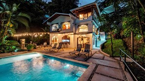 Una casa de dos pisos con piscina, zona de estar al aire libre y guirnaldas de luces.