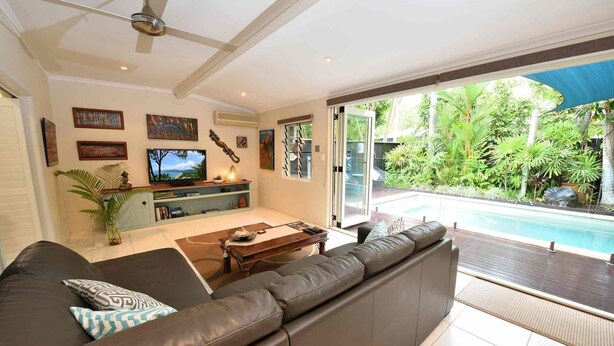 A living room with a leather sofa, a coffee table, and a TV. There is a sliding glass door leading to a pool area with a view of greenery. - Bella Leuca family getaway (Port Douglas)