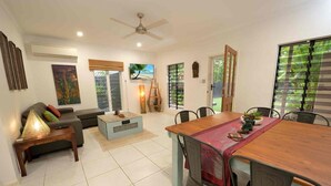 A spacious living area with a dining table, sofa, and chairs, a ceiling fan, and a view of greenery through the windows. - Bella Leuca family getaway (Port Douglas)