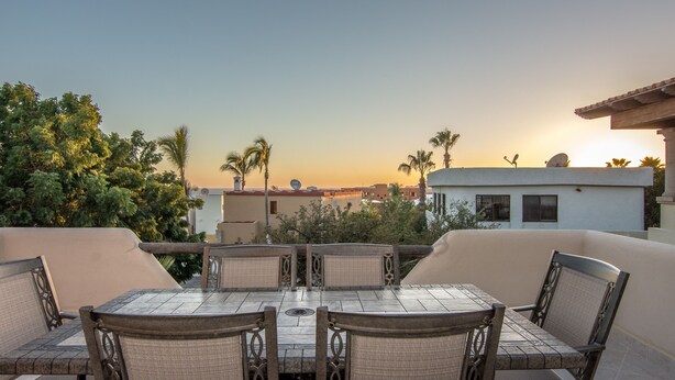 Una terraza con mesa y sillas, con vistas a una puesta de sol y palmeras.