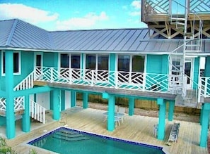 Um edifício turquesa de dois andares com piscina, área de estar ao ar livre e escadaria que leva a um terraço superior.