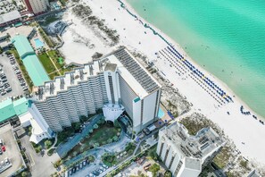 Vue aérienne d’un hôtel situé en bord de mer, avec un grand bâtiment doté d’un toit en zigzag distinctif, un espace de stationnement et une plage de sable parsemée de parasols.