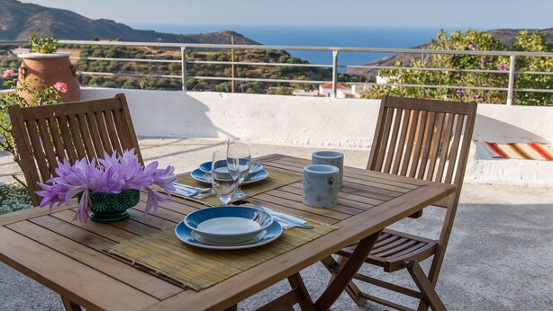 Una mesa de madera dispuesta con platos, cubiertos y un jarrón de flores moradas, con vistas a un paisaje costero.
