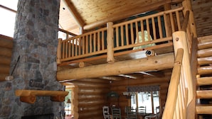 Interior de una cabaña de troncos con chimenea de piedra, balcón de madera y mobiliario rústico.