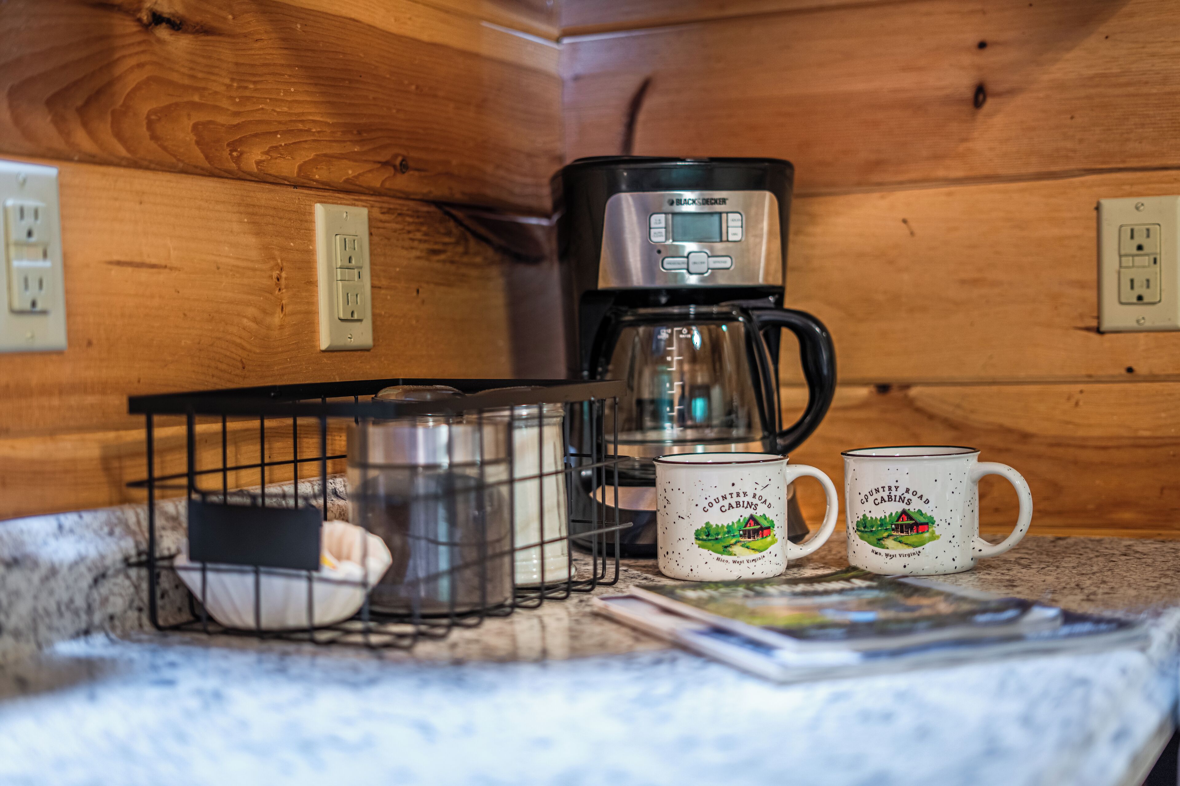 Uma cozinha com cafeteira, duas canecas, um castiçal e uma revista sobre o balcão.