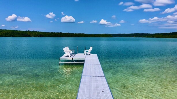 A clear lake with a dock extending into the water, featuring two white chairs and a table.