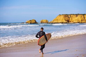 Une personne vêtue d’une combinaison de plongée, portant une planche de surf, se trouve sur une plage, avec des falaises rocheuses en arrière-plan.