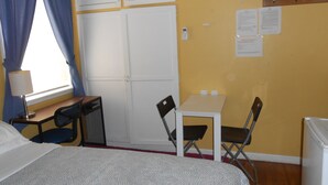 Een kamer met een bed, een bureau, een stoel, een klein tafeltje, een raam met gordijnen en een wandplank met papieren.