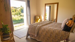 Un dormitorio con una cama grande, una silla, una mesita de noche con un florero y vistas a un jardín a través de una puerta corredera.