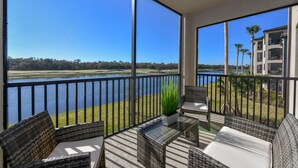 A balcony with wicker furniture, a potted plant, and a view of a lake and buildings.