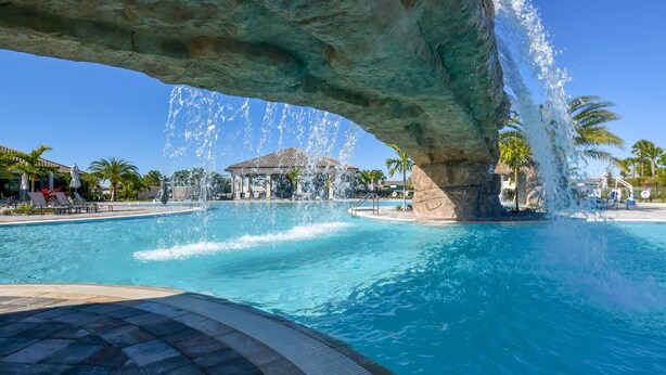 A swimming pool with a large rock arch and water fountains, surrounded by palm trees and lounge chairs.