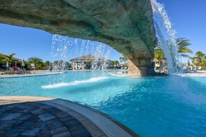 Piscina con un gran arco de roca y fuentes de agua, rodeada de palmeras y sillas de descanso.