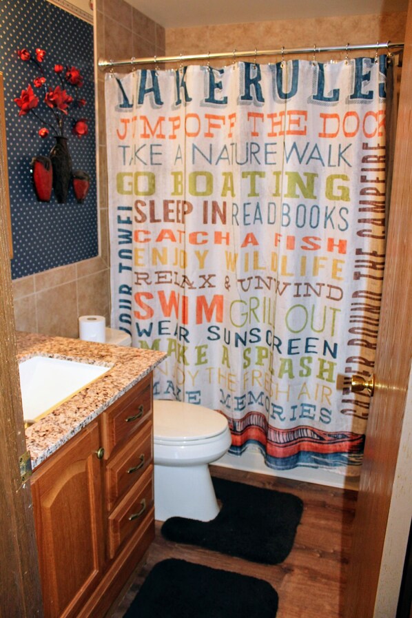 A bathroom with a shower curtain featuring various words and phrases, a toilet, a sink with a granite countertop, wooden cabinets, and a black rug on the floor.