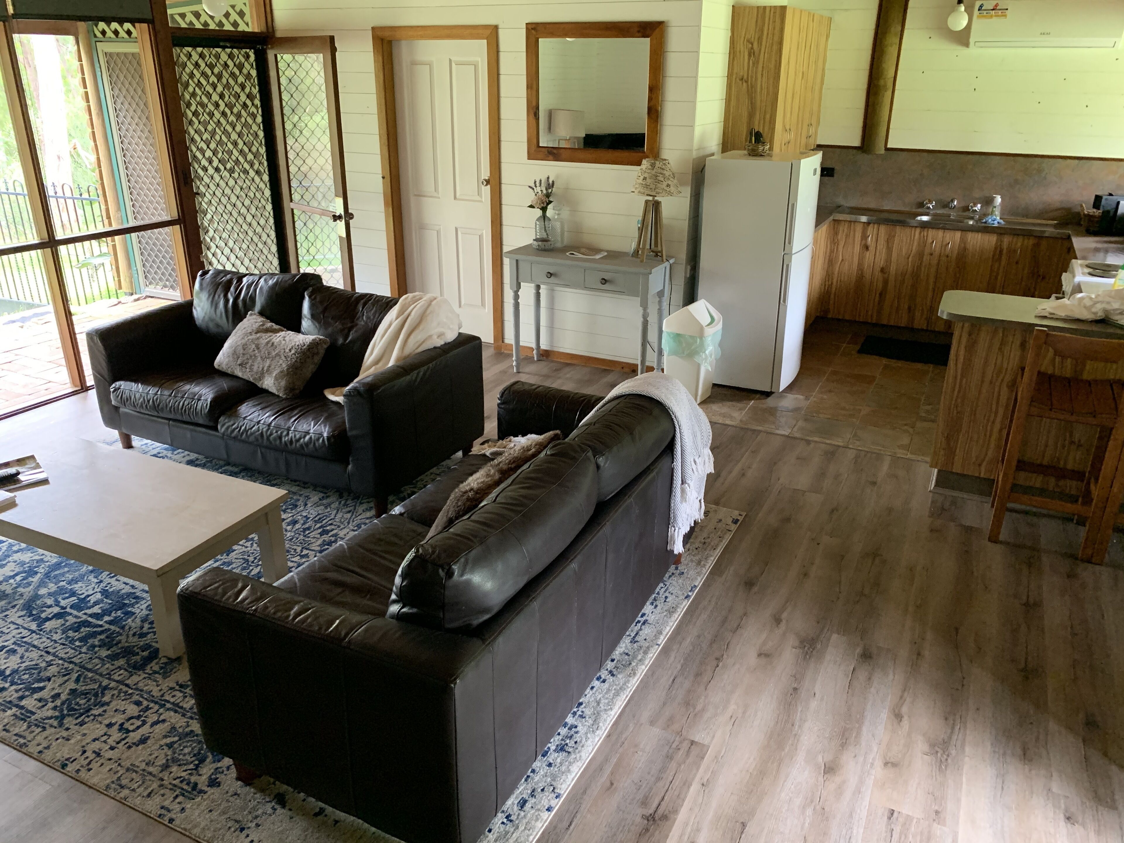 A living room with a sofa, a coffee table, and a rug. There is a kitchen area with a sink and a refrigerator.