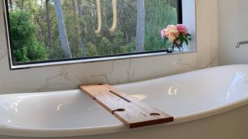Ein modernes Badezimmer mit einer freistehenden Badewanne, einem hölzernen Tablett und einem Fenster mit Blick auf Bäume.