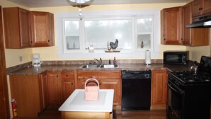 A kitchen with wooden cabinets, a white countertop, a stove, a microwave, a dishwasher, and a window with decorative items. - The Mill House, year around Country Home & Historic Grist mill w/ 2 Kayaks (Posen)